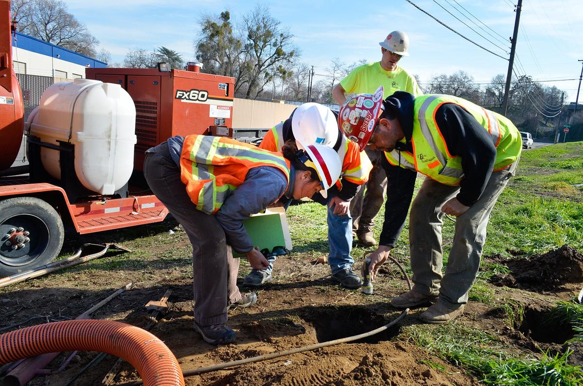 Crew in hard hats leaning over a small pothole to check an exposed utility line, a vacuum excavation unit behind them